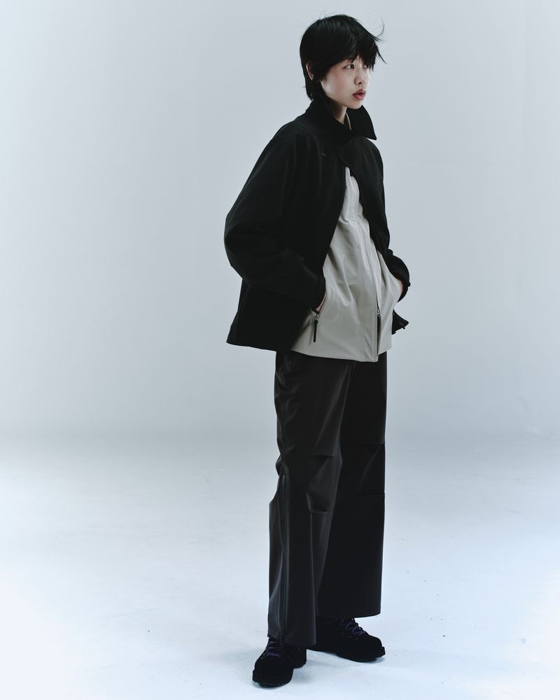 Full shot of a non-binary person posing in black jacket and loose black pants against a bright grey studio backdrop in soft lighting.