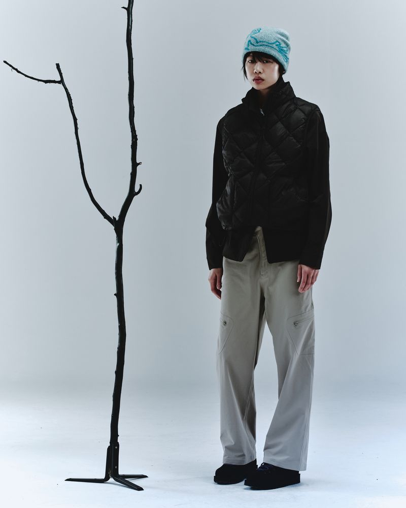 Asian model posing in black puffer vest, beige cargo pants, and blue beanie in a white studio with a minimalist tree prop.