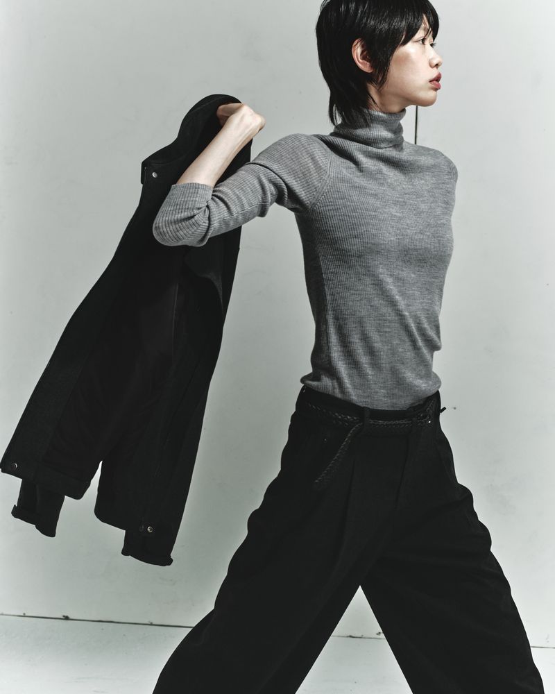Studio shot of a stylish Asian woman wearing gray turtleneck and black pants, holding a black coat over her shoulder, looking away.