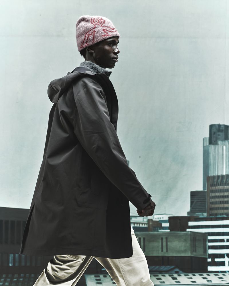 Dark-skinned adult male in coat and knit hat walking against a city backdrop with gray sky; fashion forward and moody.