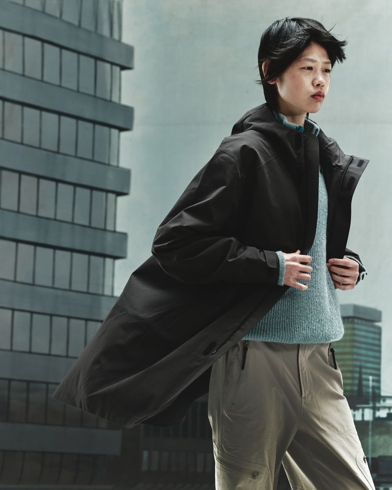 Woman poses in long dark coat, light-blue sweater, tan pants against city buildings. Fashion shot with overcast lighting.