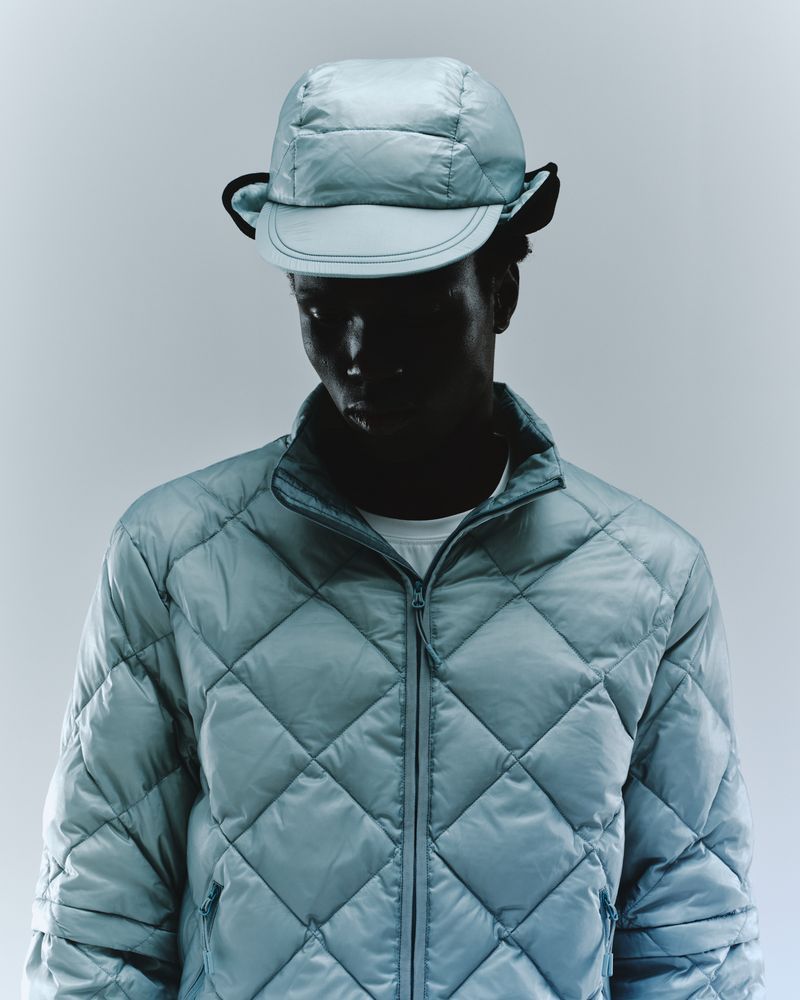 Person wearing a blue quilted jacket and matching hat, standing in front of a light background in a studio shot. Fashion concept.