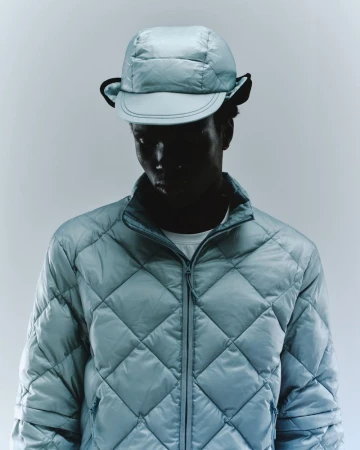Person wearing a blue quilted jacket and matching hat, standing in front of a light background in a studio shot. Fashion concept.