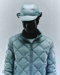 Man wearing a light blue quilted puffer jacket and matching padded cap against a seamless gradient background.