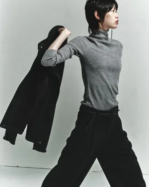 Model with dark hair wearing a grey turtleneck and black trousers holding a jacket over her shoulder against a plain white studio wall