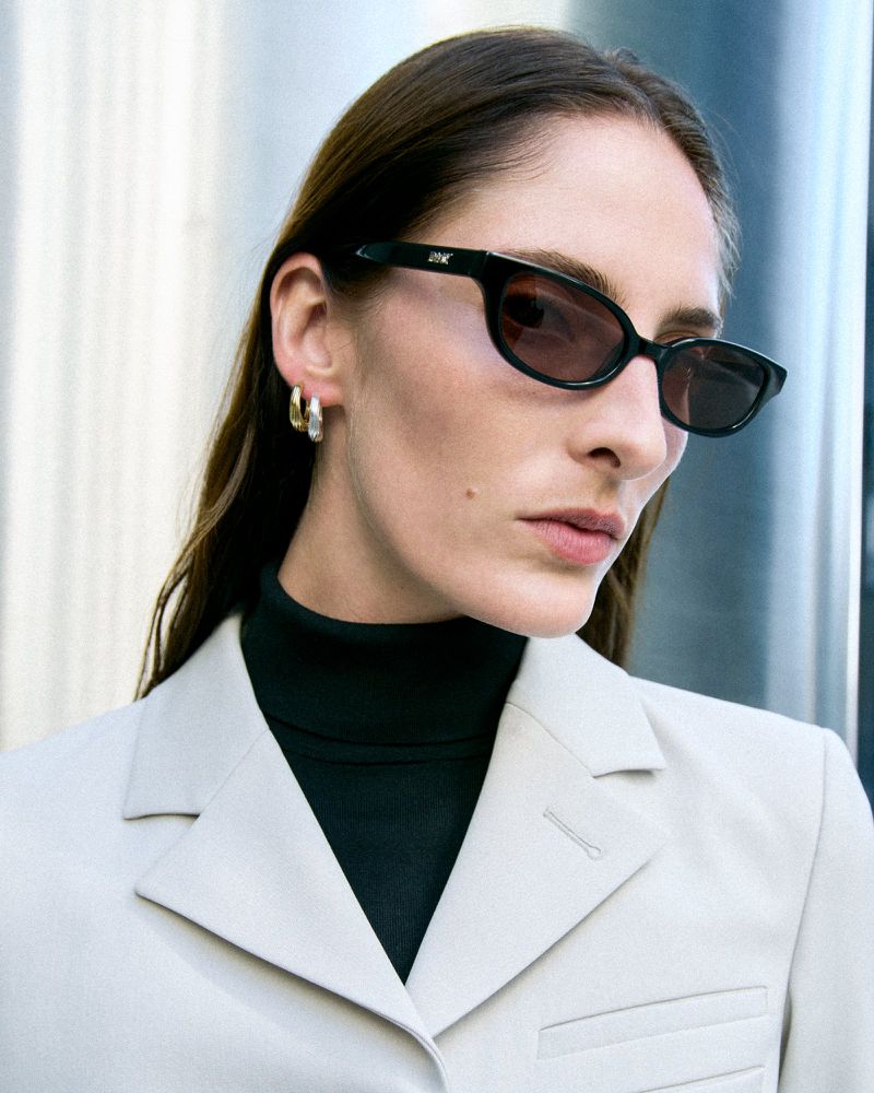 Model Annie Tice wearing black oval sunglasses, a cream tailored blazer, and double hoop earrings.