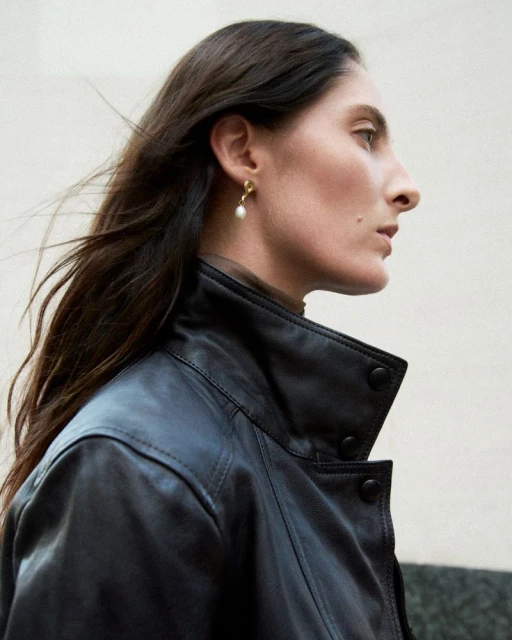 A woman in a black leather jacket poses in a side-view shot. Her dark hair is windswept and she wears a gold earring with a pearl.