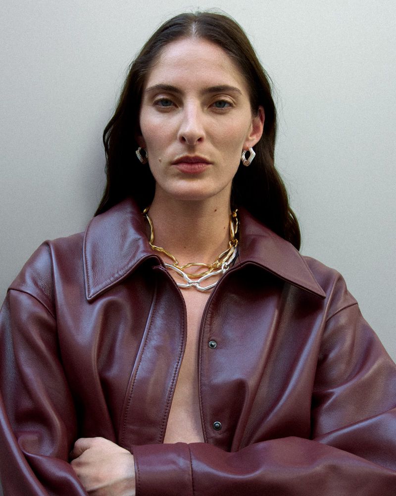 Brunette woman in a burgundy leather jacket and chunky jewelry poses indoors against a neutral wall in a medium studio shot.