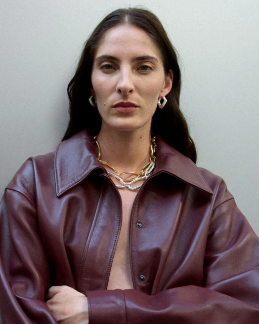 Brunette woman in a burgundy leather jacket and chunky jewelry poses indoors against a neutral wall in a medium studio shot.