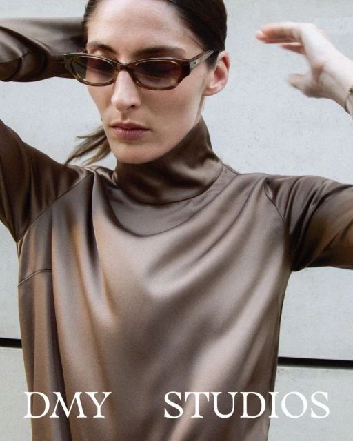 Woman wearing sunglasses and a satin turtleneck top poses indoors with hands near her head in a fashion or beauty editorial image.