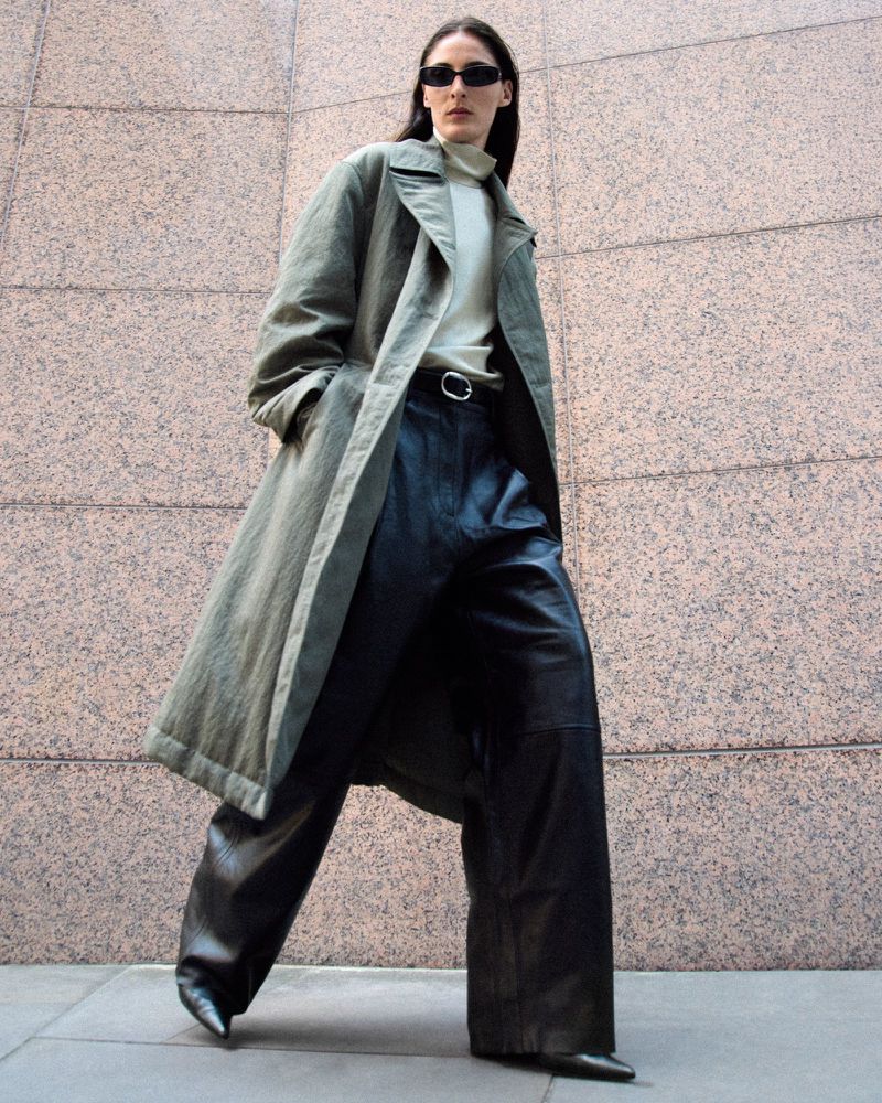 Model wearing dark sunglasses, an olive trench coat, and dark leather trousers standing before stone exterior walls.