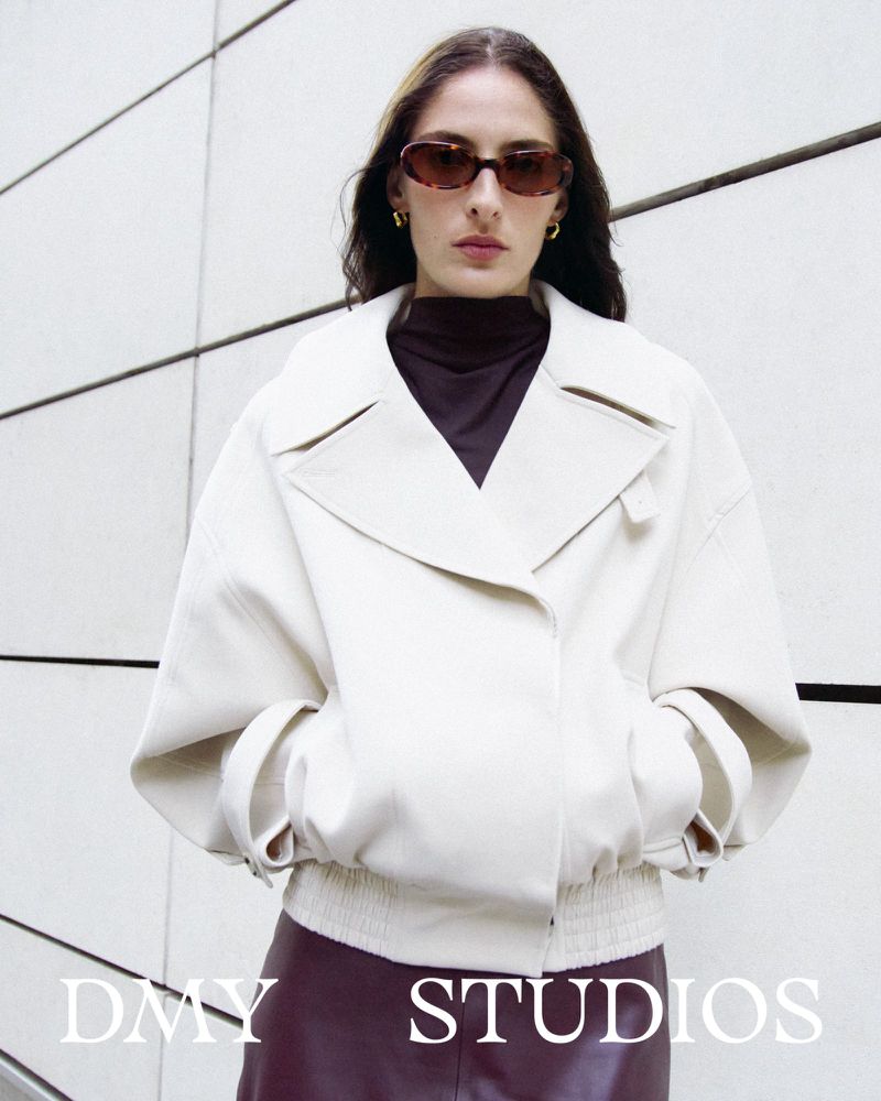 A fashionable woman in an off-white jacket poses against a geometric wall, showcasing style with sunglasses and gold earrings in an urban setting.