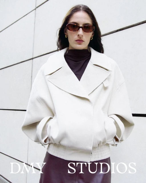 A fashionable woman in an off-white jacket poses against a geometric wall, showcasing style with sunglasses and gold earrings in an urban setting.