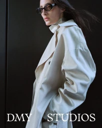 Model Annie Tice wearing dark oval sunglasses and an oversized cream trench coat against a black background.