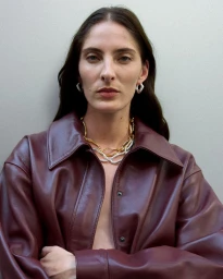 Model Annie Tice wearing a burgundy leather jacket with layered gold and silver chain necklaces against a neutral studio backdrop.