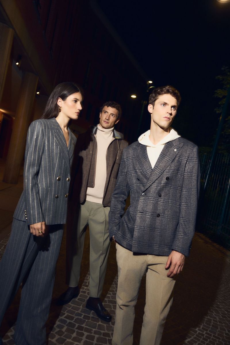 Three fashionably dressed people stand outside in the evening, showcasing modern blazers and stylish, tailored pants.