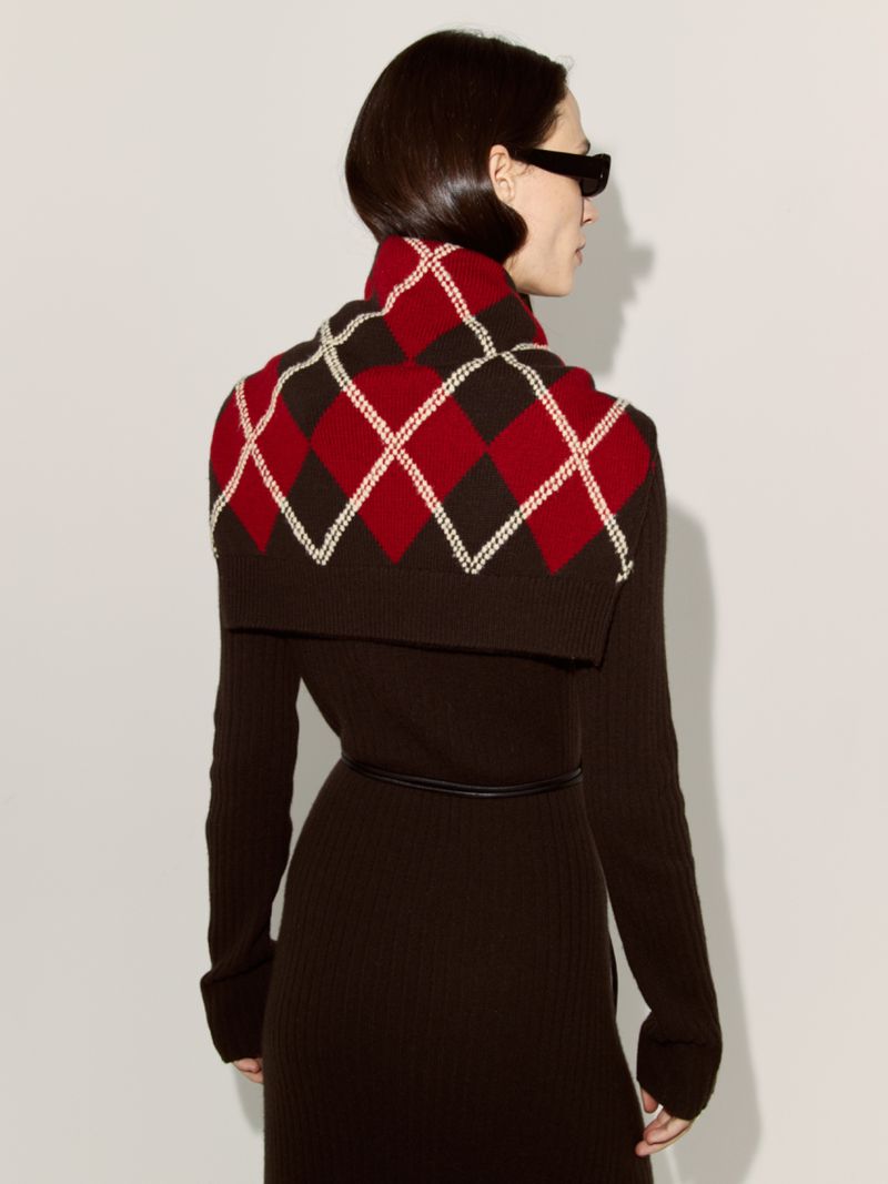 Woman in brown dress with red and brown argyle shrug against white background, looking away, fashion shot.