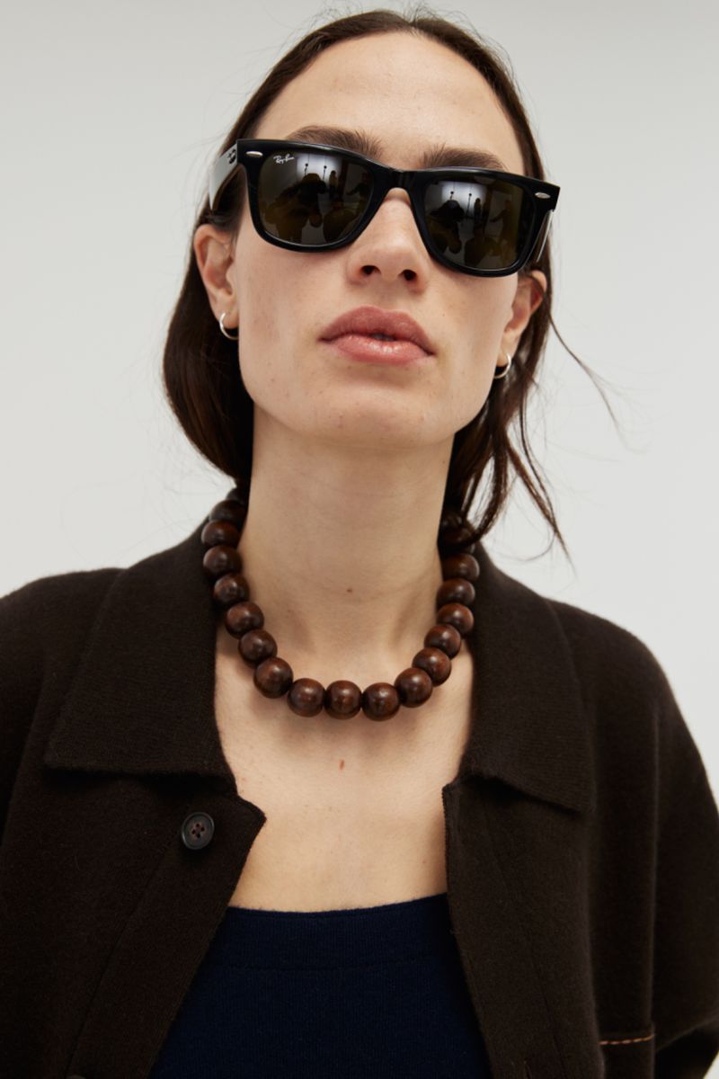 Close-up of a woman with long brown hair wearing black sunglasses, a brown beaded necklace, and a brown jacket against white.