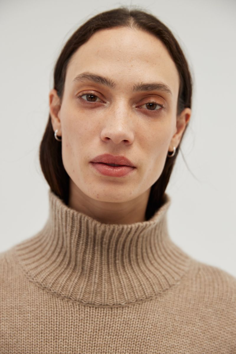 Close-up portrait of a light-skinned adult woman with neutral expression, wearing a tan turtleneck sweater against a plain white background.