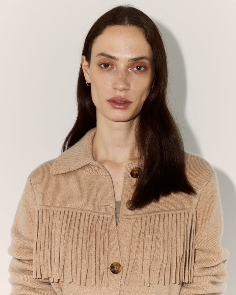 Portrait of a brunette adult woman wearing a tan fringe jacket indoors against a light-colored wall, medium shot, eye-level angle.