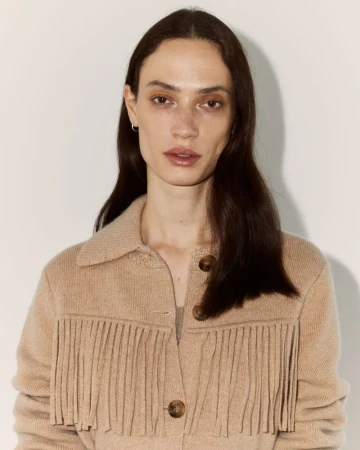Portrait of a brunette adult woman wearing a tan fringe jacket indoors against a light-colored wall, medium shot, eye-level angle.