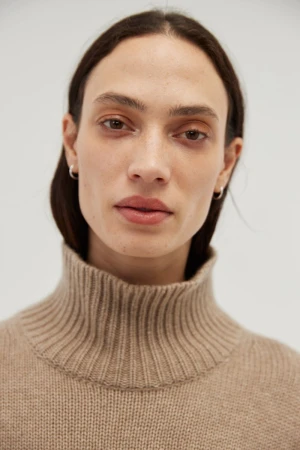 Model Sophie Koella wearing a beige ribbed turtleneck knit sweater against a light grey studio background.
