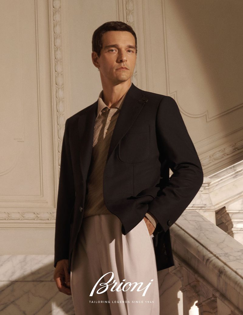 A sharply dressed man in a dark blazer and light trousers stands on a marble staircase in a luxurious, naturally lit setting.
