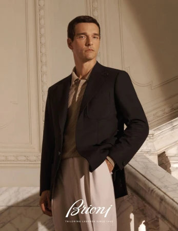 A sharply dressed man in a dark blazer and light trousers stands on a marble staircase in a luxurious, naturally lit setting.