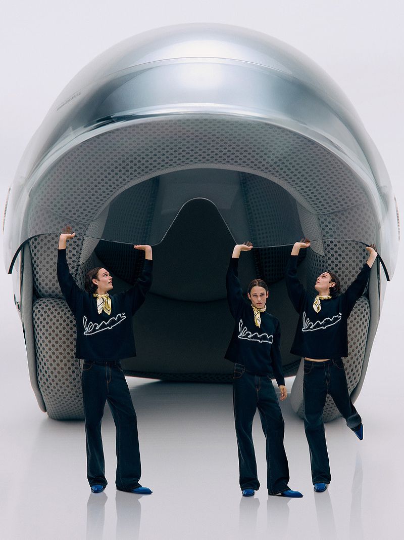 Three adults in blue outfits support a huge motorcycle helmet in a studio setting, creating a surreal and playful fashion composition.