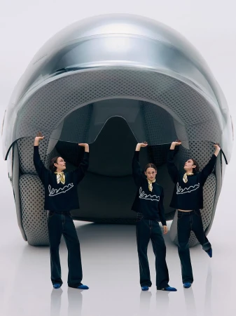 Three adults in blue outfits support a huge motorcycle helmet in a studio setting, creating a surreal and playful fashion composition.