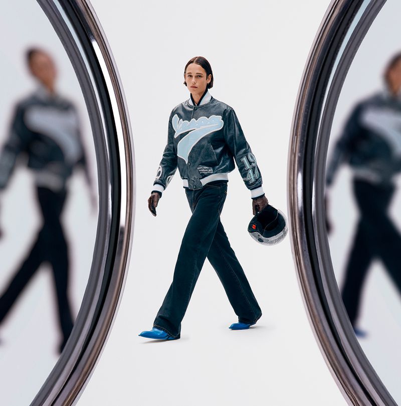 A full shot of a person walking toward the camera, wearing a bomber jacket, dark pants, and blue shoes, holding a helmet.