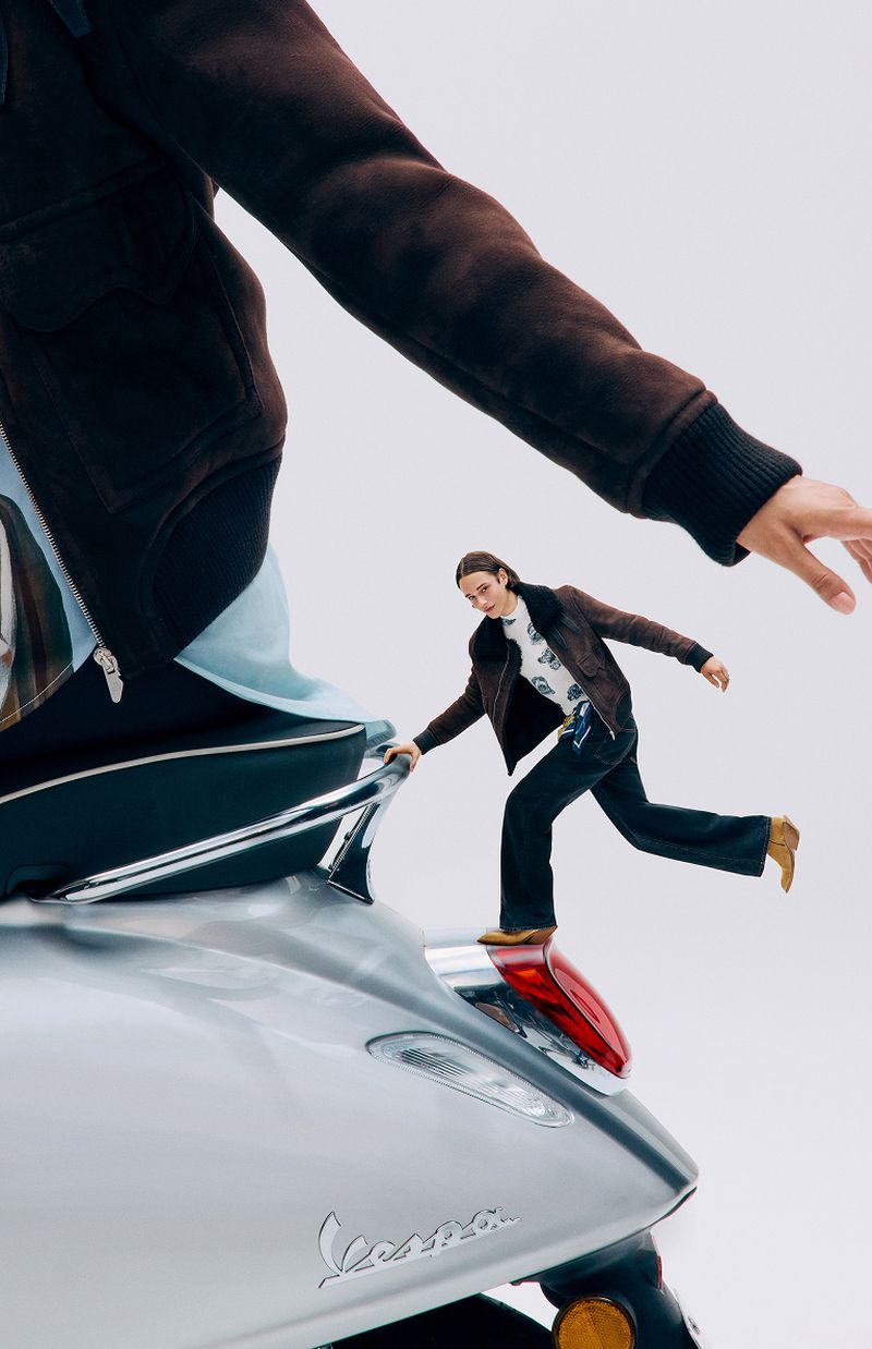 Person balancing on a silver Vespa scooter in a studio setting, wearing a brown jacket and dark pants, captured in a fashion editorial style.