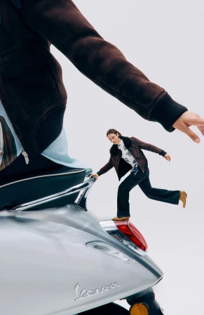 Person balancing on a silver Vespa scooter in a studio setting, wearing a brown jacket and dark pants, captured in a fashion editorial style.