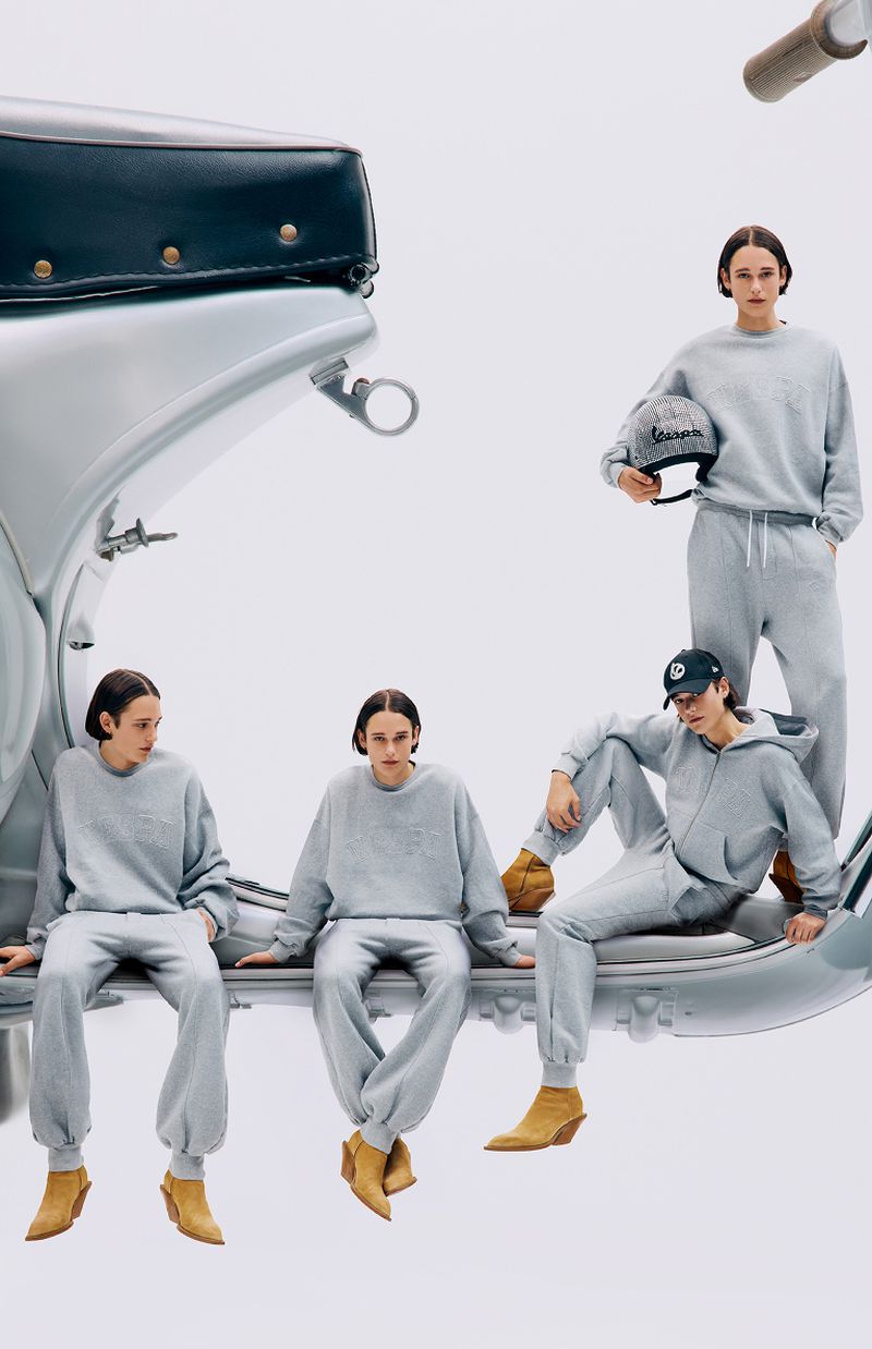 A group of four adult models wearing gray sweatsuits, posing with a silver Vespa scooter in a brightly lit photo studio setting.