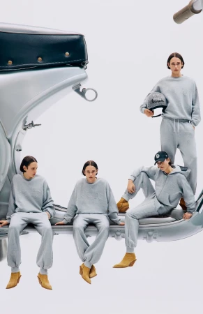A group of four adult models wearing gray sweatsuits, posing with a silver Vespa scooter in a brightly lit photo studio setting.
