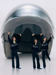 Models in black logo sweatshirts pose holding the visor of a giant metallic silver Vespa helmet in a white studio.