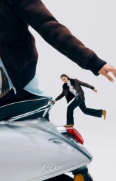 A model balances on the rear light of a silver Vespa scooter against a white seamless background with a foreground arm reaching in.