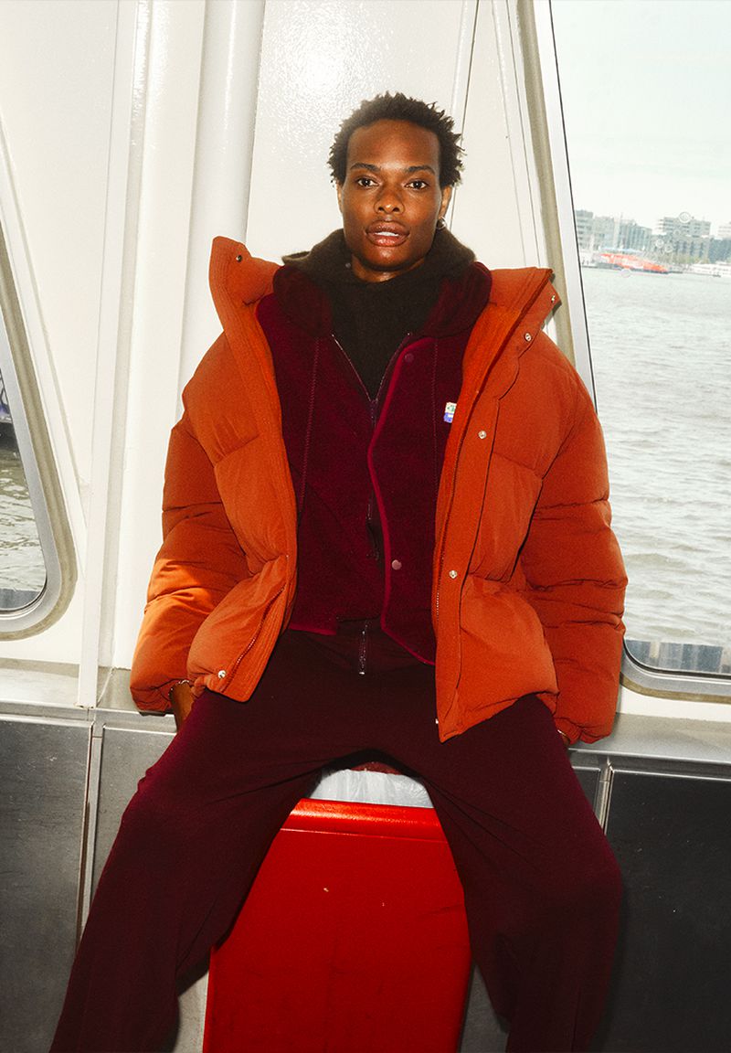 A stylish Black male model poses indoors near a window, wearing an orange puffer jacket over a burgundy outfit, with city scenery outside.