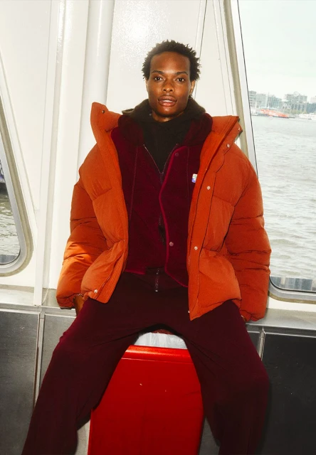A stylish Black male model poses indoors near a window, wearing an orange puffer jacket over a burgundy outfit, with city scenery outside.
