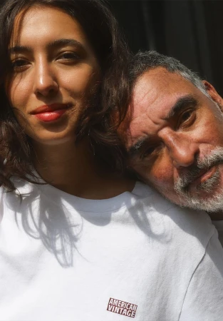 Close-up portrait of a young woman in a white t-shirt with an older man's head resting on her shoulder in soft light.