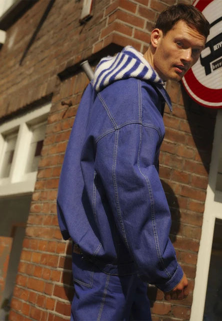 An adult man in a denim jacket and striped hoodie is captured in an eye-level, medium shot in an urban setting with a brick background.