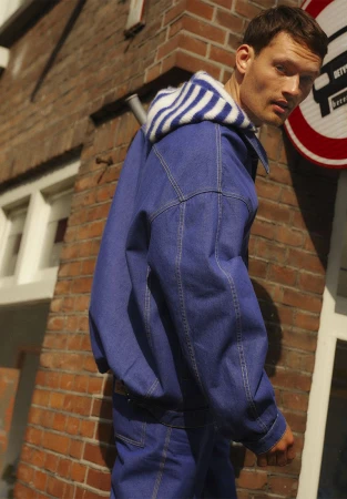 An adult man in a denim jacket and striped hoodie is captured in an eye-level, medium shot in an urban setting with a brick background.