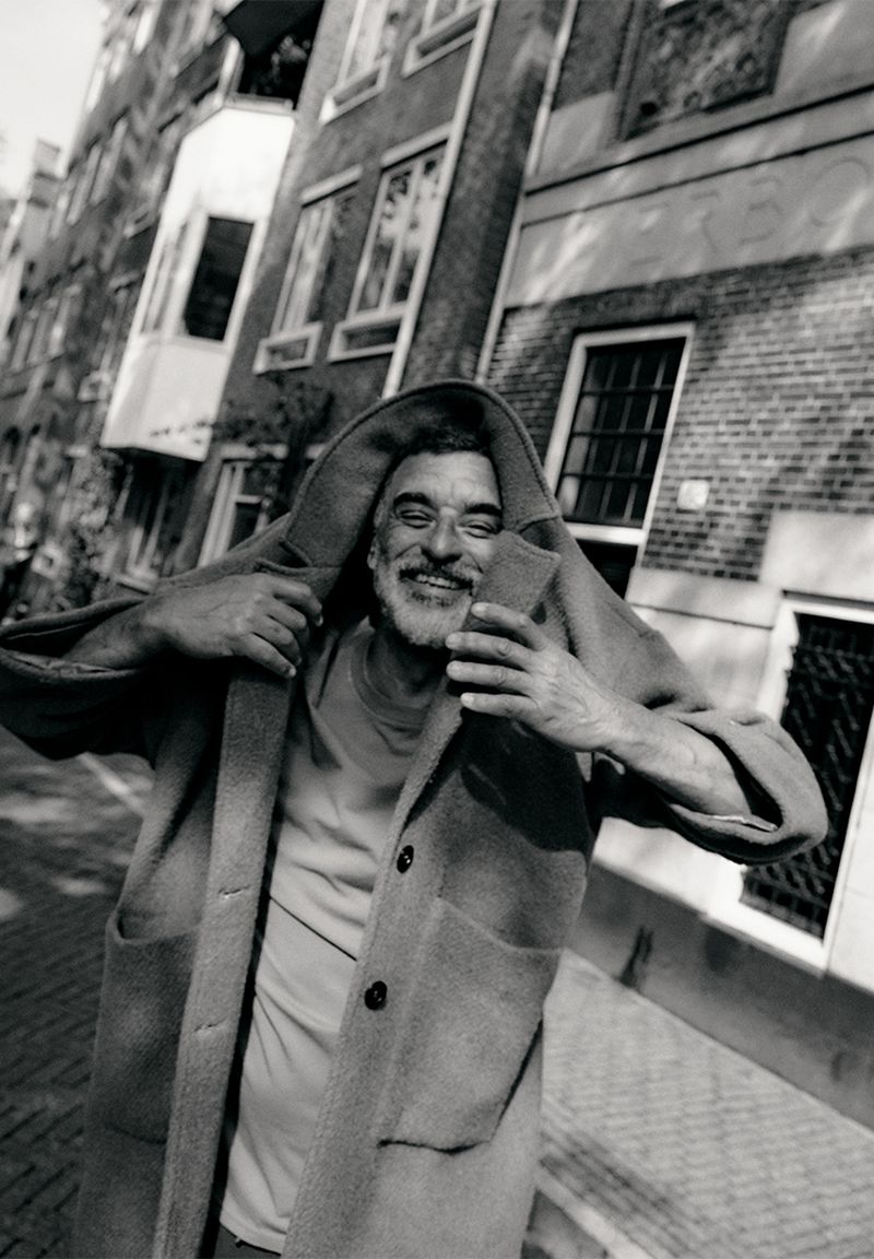 Smiling older man in Amsterdam wearing a hooded coat, photographed by Rasmus Weng Karlsen in black and white.