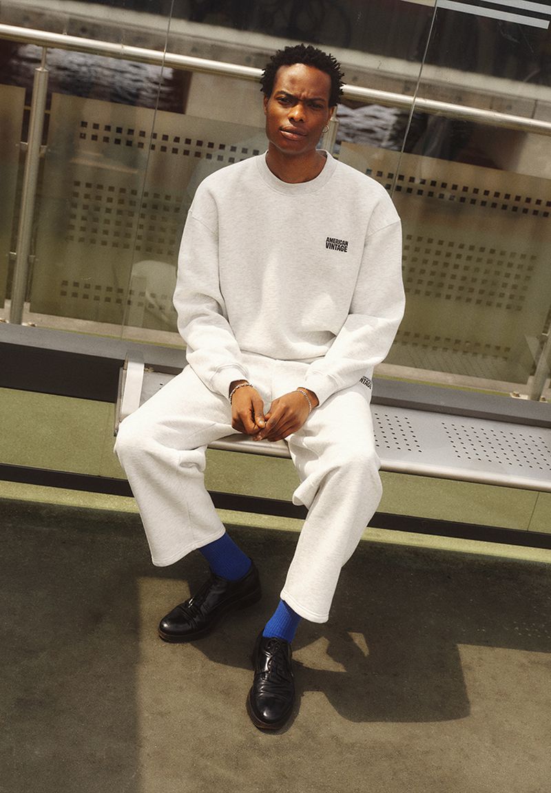 An adult male sits on a bench wearing a grey sweatshirt and pants, blue socks, and black dress shoes in a neutral indoor environment.