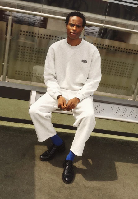 An adult male sits on a bench wearing a grey sweatshirt and pants, blue socks, and black dress shoes in a neutral indoor environment.