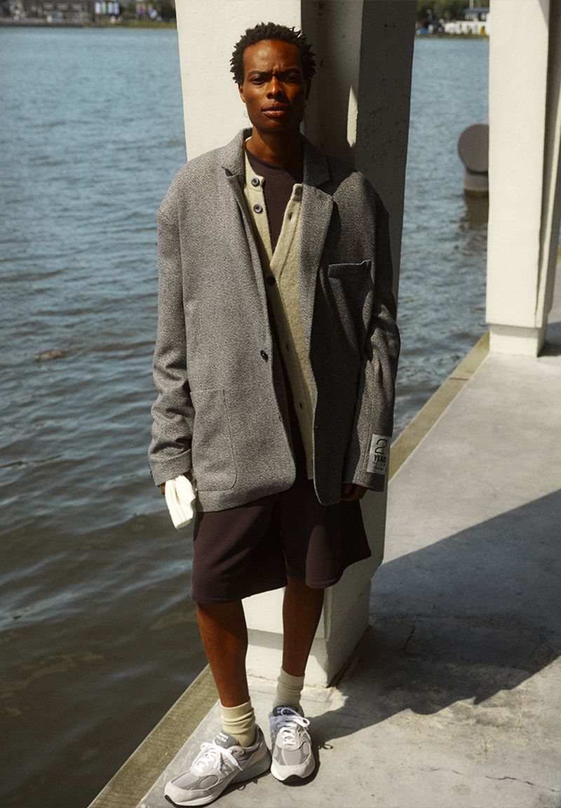 Adult male poses in blazer and shorts in an urban waterfront setting, showcasing fashion and lifestyle photography with natural lighting.