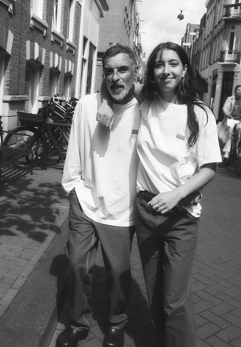 Black and white medium shot of an adult man and a young adult woman standing arm-in-arm on a city street with bicycles parked in the background.