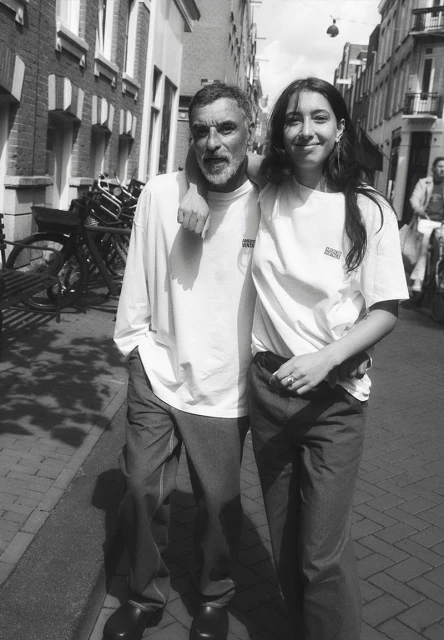 Black and white medium shot of an adult man and a young adult woman standing arm-in-arm on a city street with bicycles parked in the background.