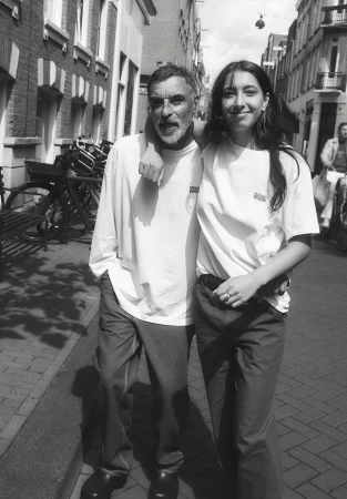 Black and white medium shot of an adult man and a young adult woman standing arm-in-arm on a city street with bicycles parked in the background.