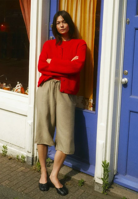 Woman in red sweater and culottes standing in doorway with yellow curtain. Relaxed, casual fashion in outdoor setting.